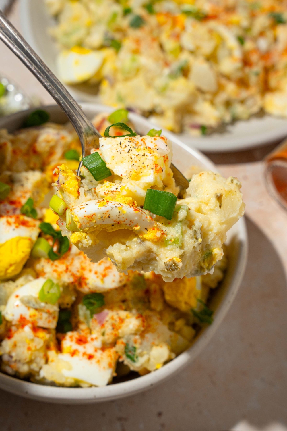 A close up of a fork with a bite of deviled egg potato salad garnished with sliced green onion and paprika. There is a bowl of potato salad on a tan counter blurred in the background.
