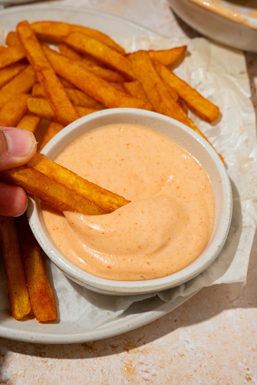 A bowl of french fry sauce on a plate with fries lined with parchment paper. Fries are being dipped into the sauce. The plate is on a tan counter.