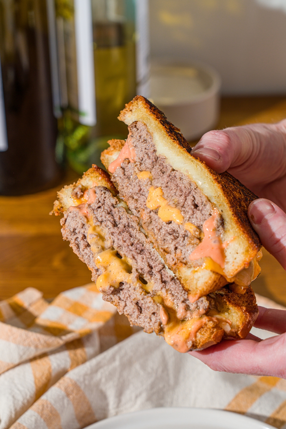 A hand holding a frisco melt with frisco sauce sliced in half and stacked on top of one another. There is a wooden counter with a white striped napkin in the background.