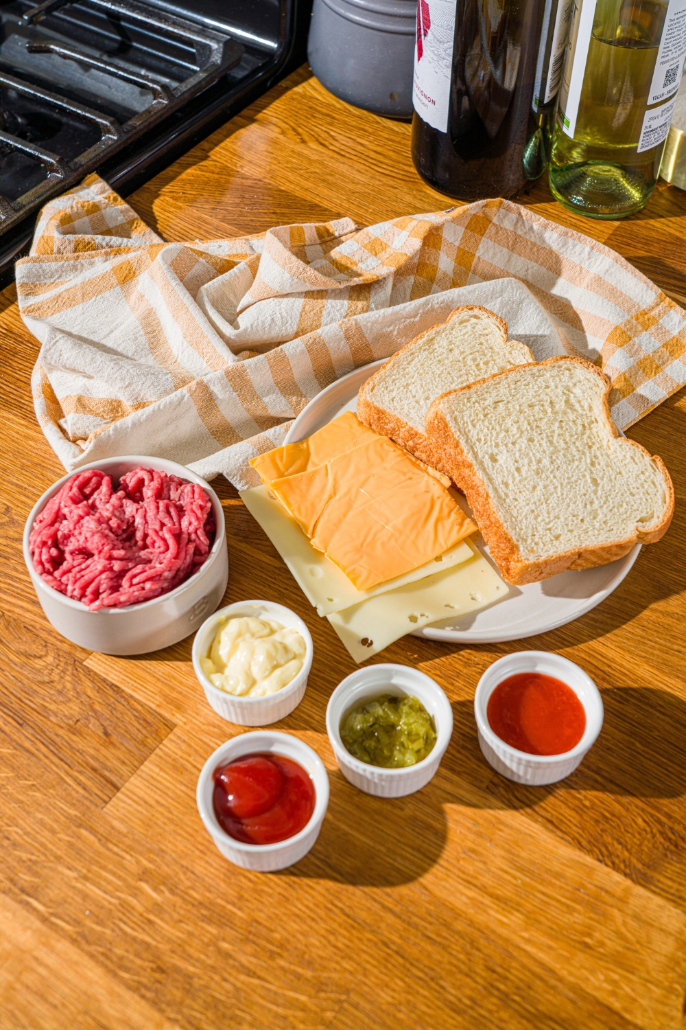 A wooden counter with several bowls in various sizes containing ingredients to make frisco melt with frisco sauce including ground beef, bread, slices of cheese, mayo, ketchup, relish, and sriracha.