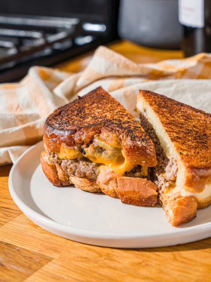 A white plate with a frisco melt with frisco sauce sliced in half. The plate is on a wooden counter with a white striped napkin.