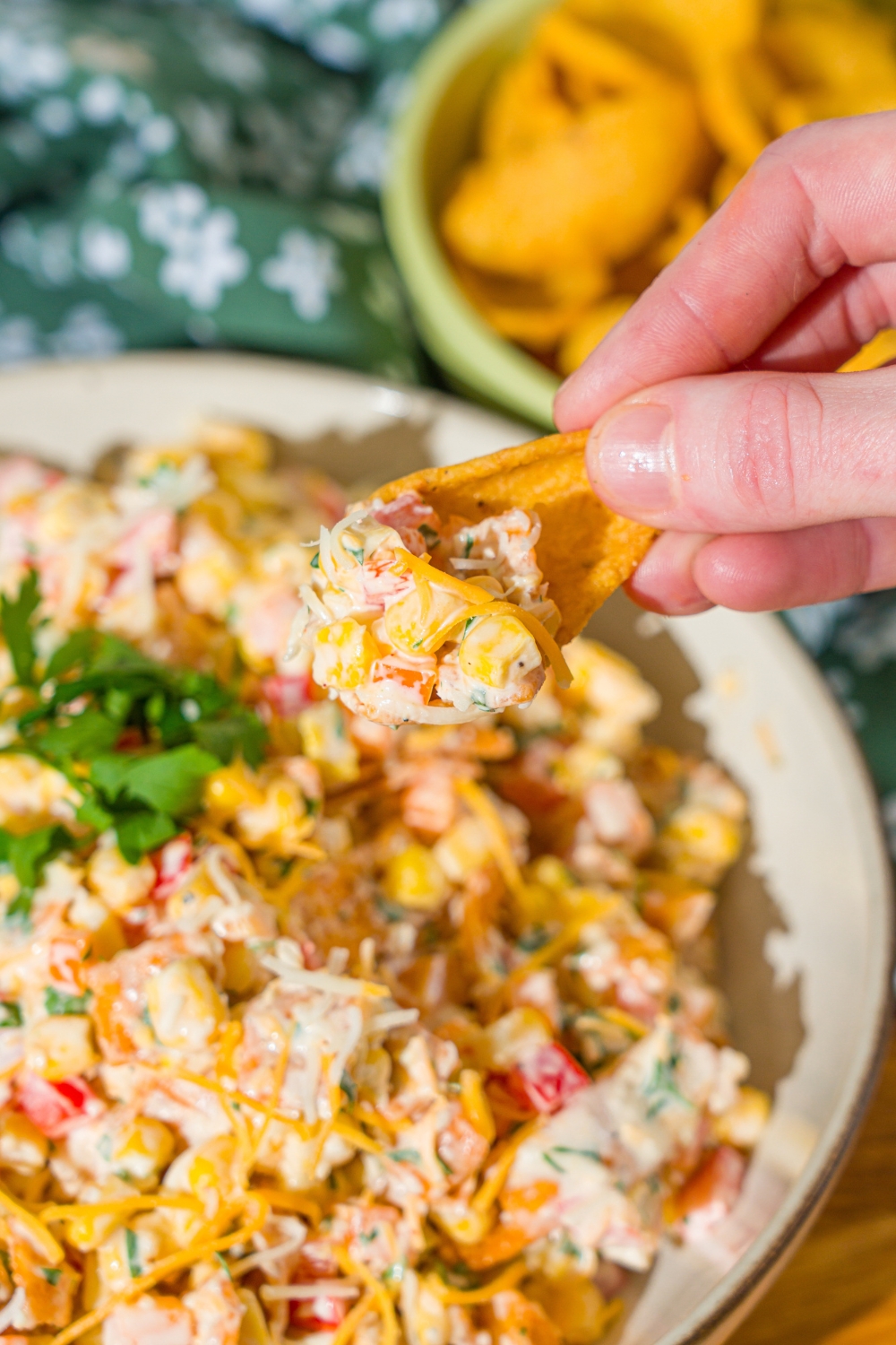 A close up of a frito chip dipped in corn salad with chili cheese fritos. There is a white bowl with frito corn salad blurred in the background.