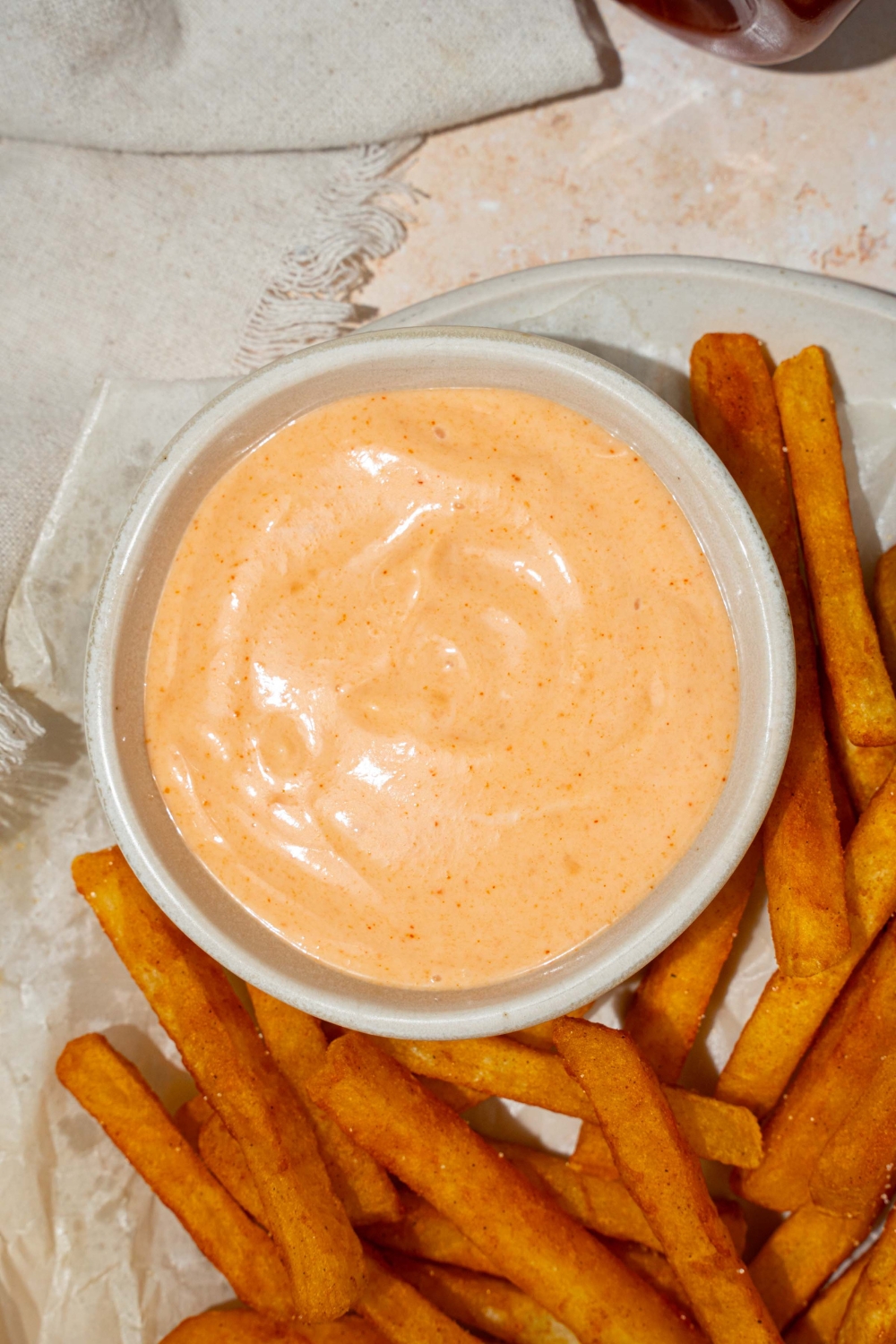 A bowl of french fry sauce on a plate with fries lined with parchment paper. The plate is on a tan counter with a white cloth napkin.