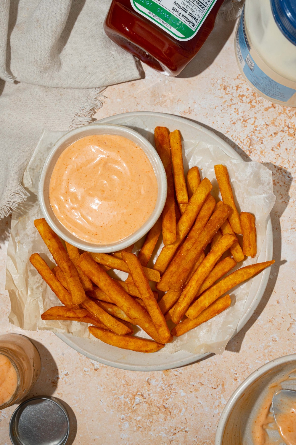 A bowl of french fry sauce on a plate with fries lined with parchment paper. The plate is on a tan counter with white cloth napkin.