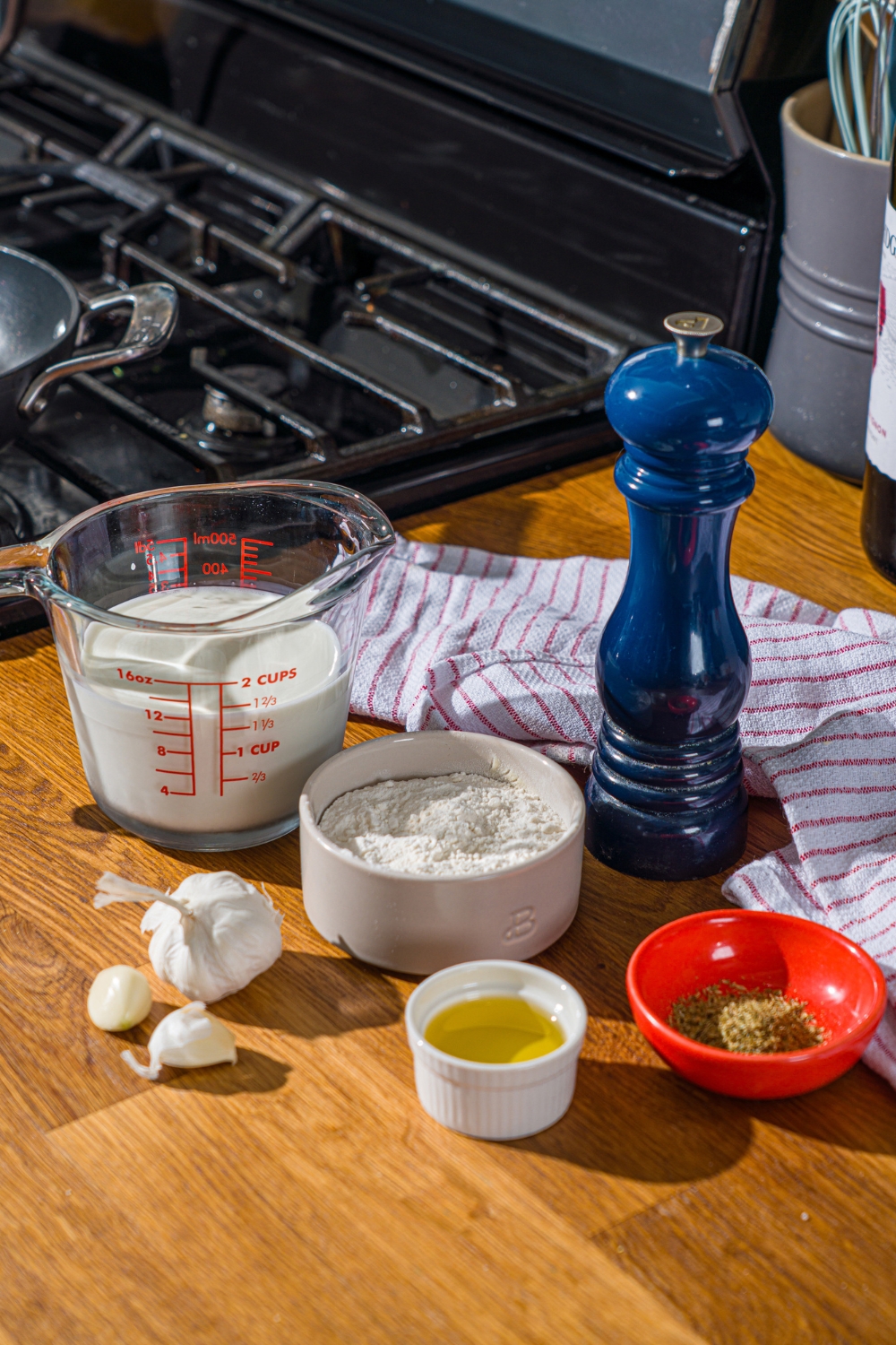 A wooden counter with ingredients to make garlic white pizza sauce including garlic, flour, oil, half and half, and seasonings.