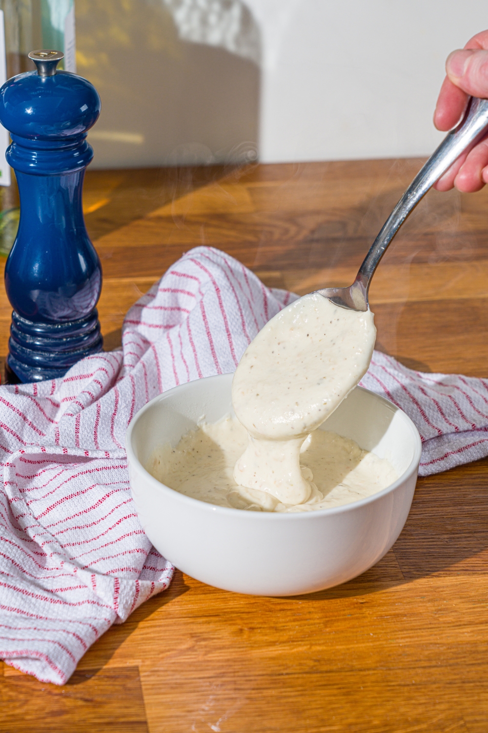 A white bowl with garlic white pizza sauce with a spoon dipping in the sauce. The bowl is on a wooden counter with a white striped napkin.