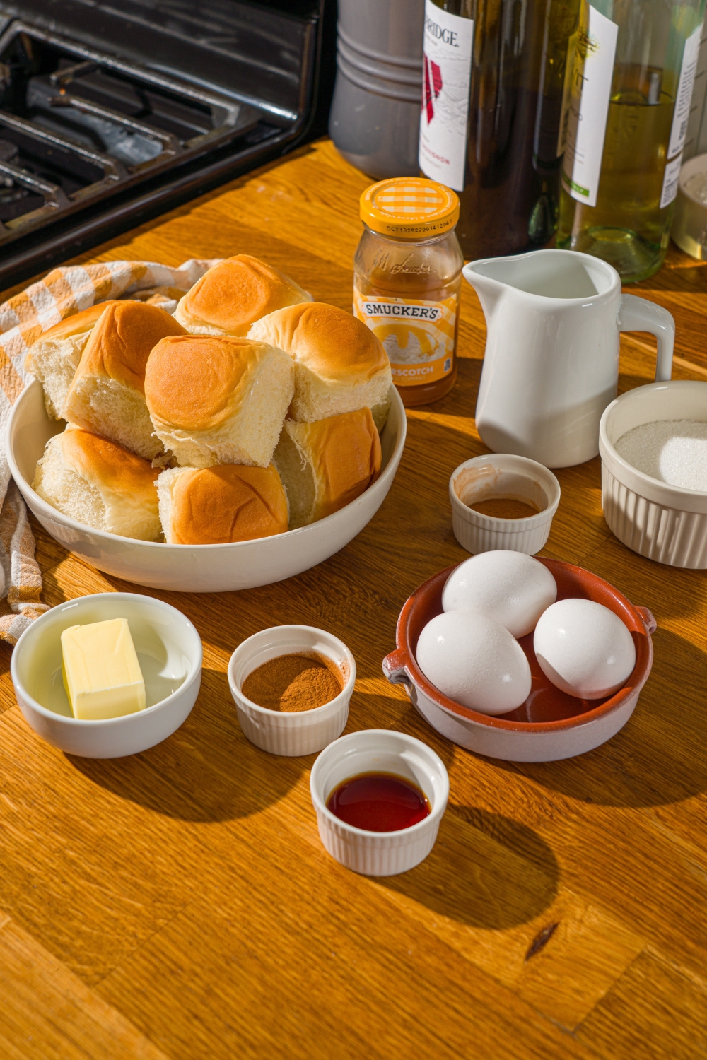 A wooden counter with several bowls in various sizes containing ingredients to make Hawaiian rolls french toast including Hawaiian rolls, half and half, milk, eggs, cinnamon, sugar, vanilla, and butter.