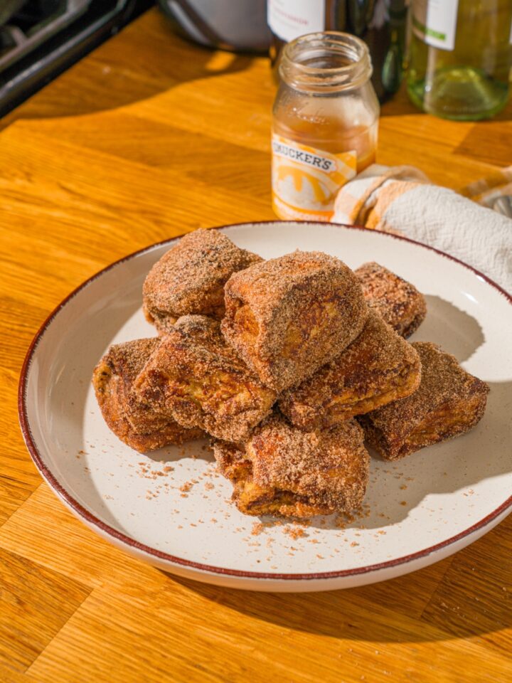 A white plate with a pile of Hawaiian roll french toast bites coated in cinnamon sugar. The plate is on a wooden counter with a yellow cloth napkin.