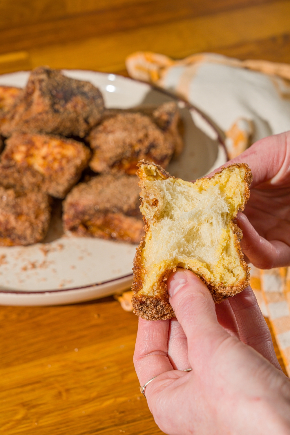 A close up of hands tearing a Hawaiian roll french toast bite in half. There is a plate of Hawaiian roll french toast bites blurred in the background.