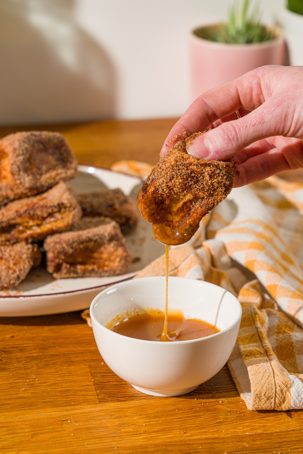 A hand dipping a Hawaiian roll french toast bite into a bowl of caramel dipping sauce. The bowl is on a wooden counter with a yellow napkin and plate of Hawaiian rolls french toast.