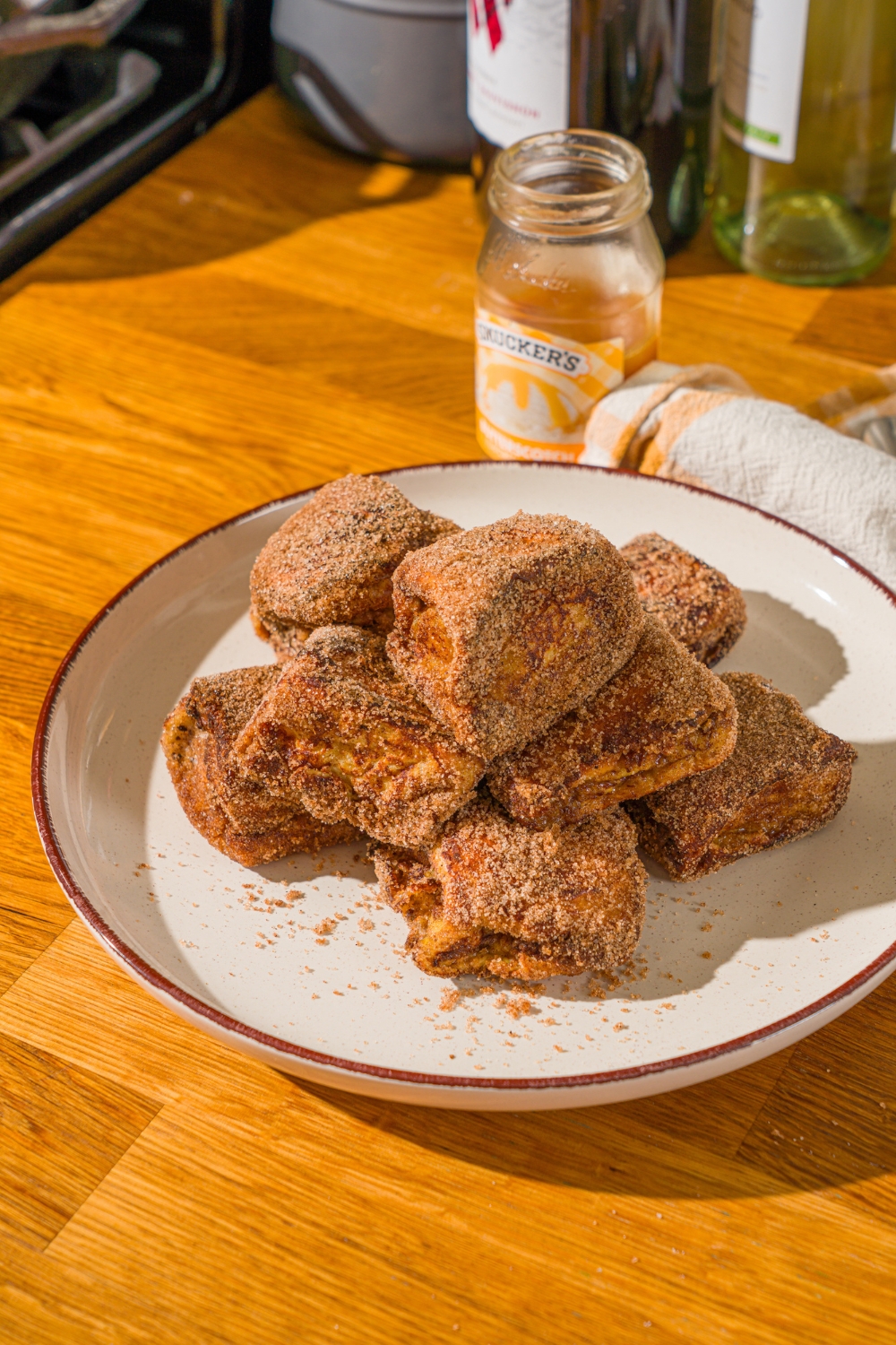 A white plate with a pile of Hawaiian roll french toast bites coated in cinnamon sugar. The plate is on a wooden counter with a yellow cloth napkin.