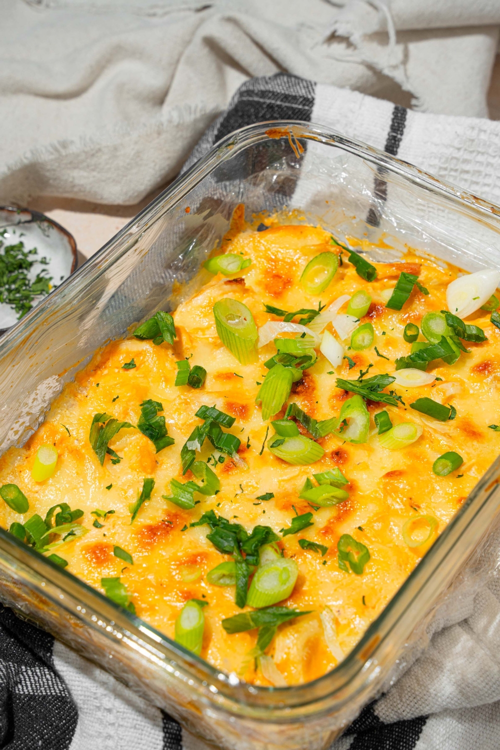 A glass baking dish with cottage cheese Buffalo chicken dip garnished with sliced green onion. The dish is on a dish towel on a tan counter with a small plate of garnishes.