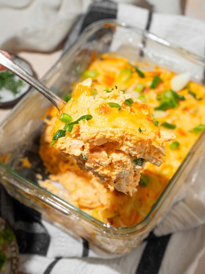 A close up of a spoon with a bite of cottage cheese Buffalo chicken dip with a baking dish of dip blurred in the background.