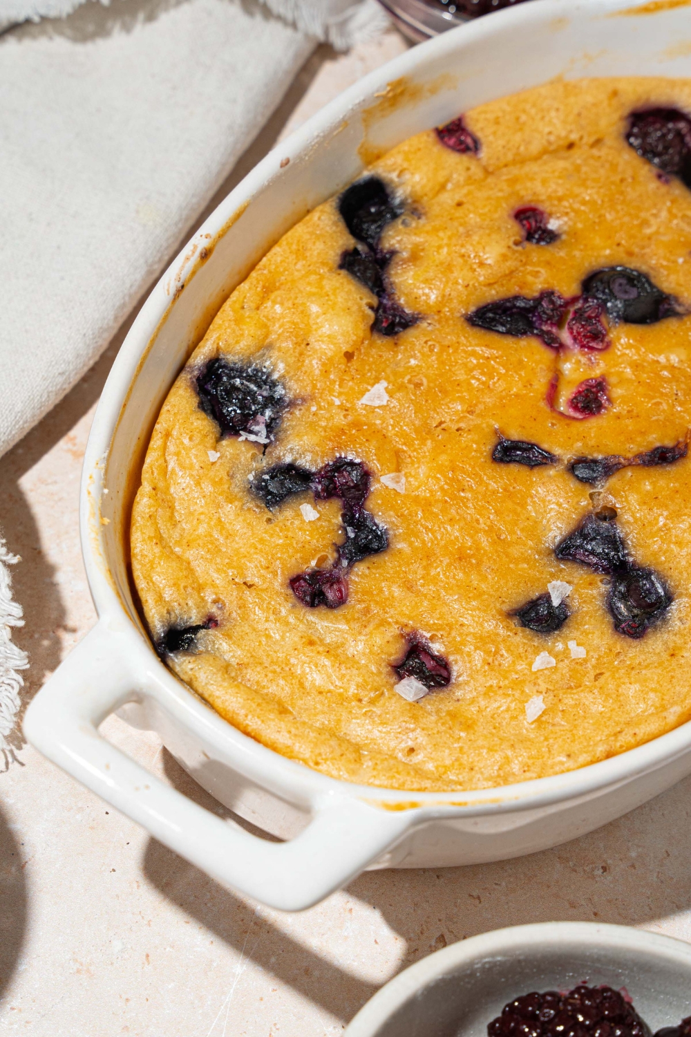 A baked protein pancake bowl with blueberries. The bowl is on a tan counter with a white cloth napkin and small bow of fruit.