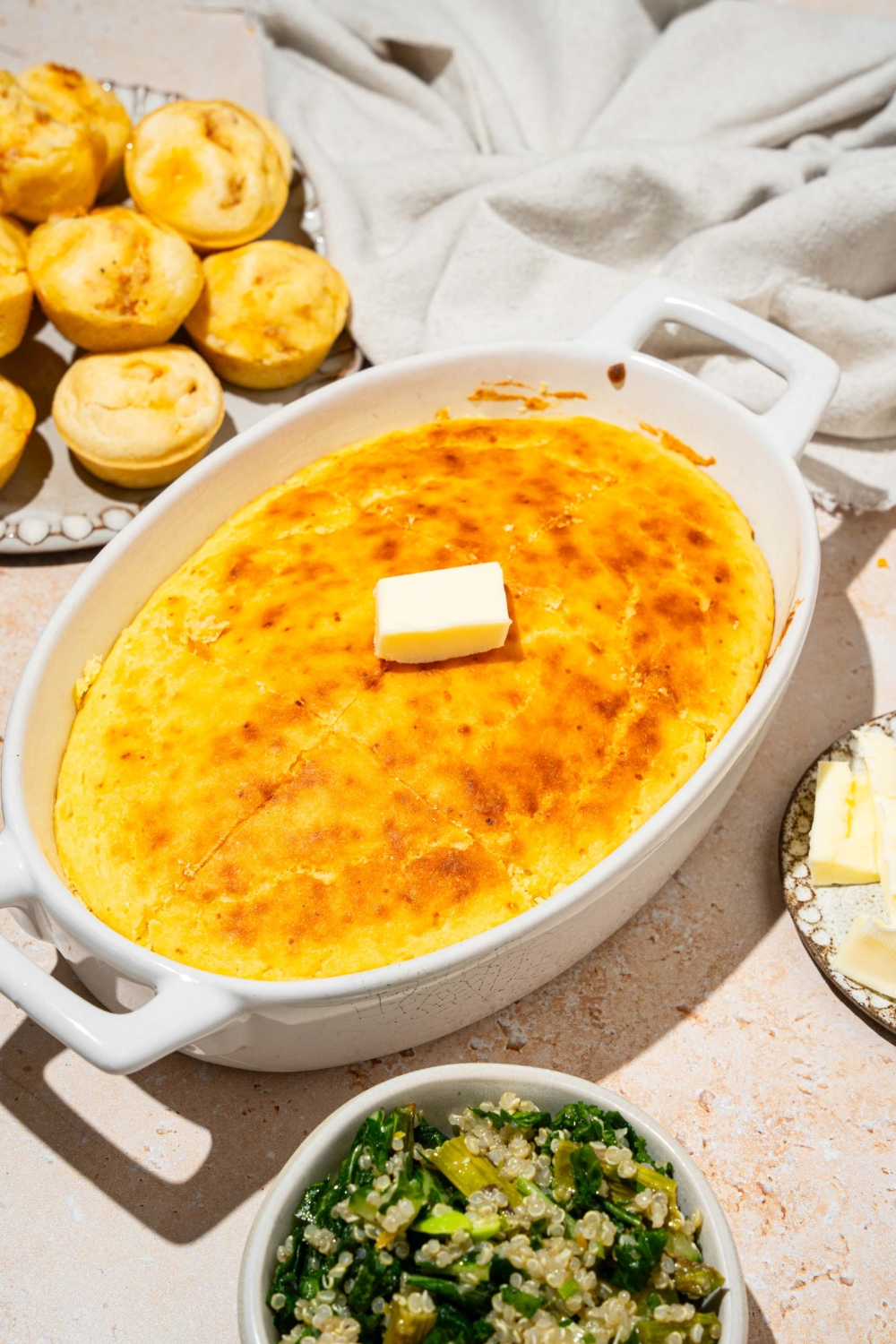 An oval baking dish with baked cornbread without eggs topped with a pat of butter. The dish is on a tan counter with a plate of muffins and white cloth napkin.
