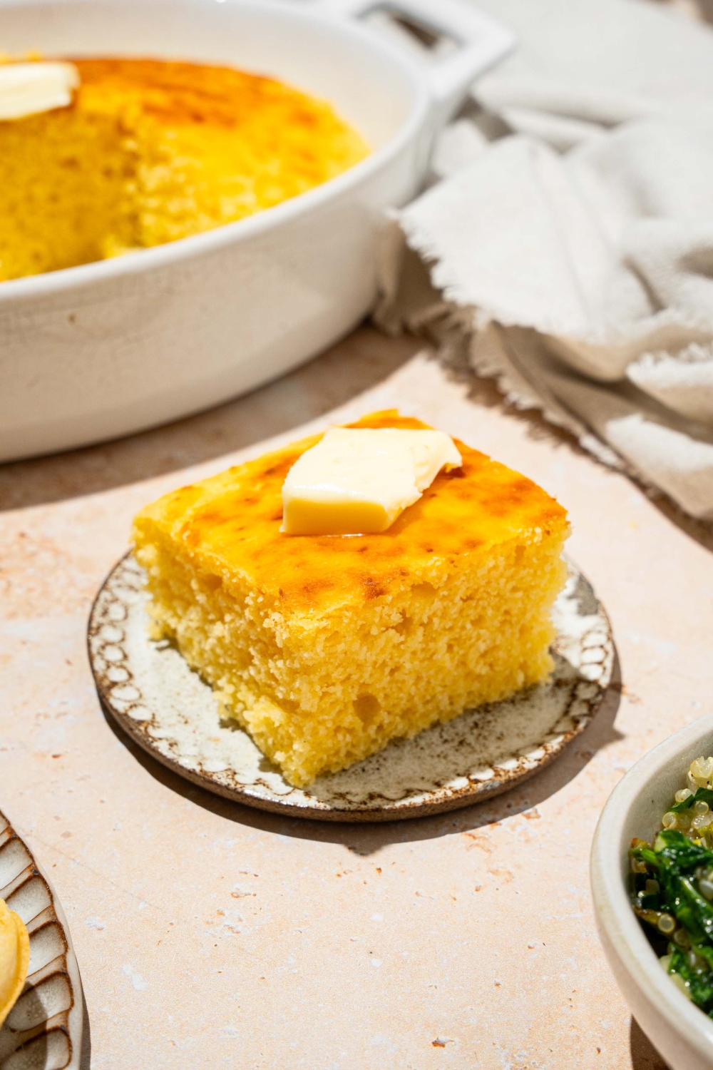 A plate with a slice of cornbread without eggs topped with a pat of butter. The plate is on a tan counter with a baking dish of cornbread and white cloth napkin.