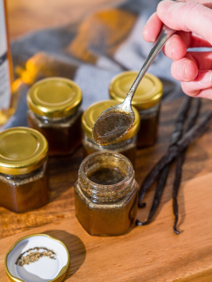 A wooden board with several jars of homemade vanilla bean paste and whole vanilla beans. A spoon is pouring the paste into one of the open jars. The board is on a wooden counter.