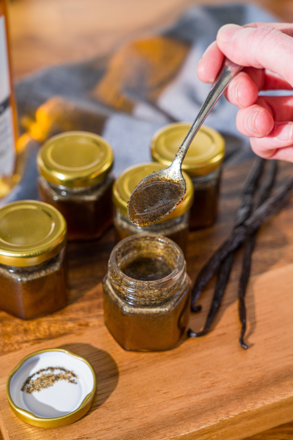 A wooden board with several jars of homemade vanilla bean paste and whole vanilla beans. A spoon is pouring the paste into one of the open jars. The board is on a wooden counter.