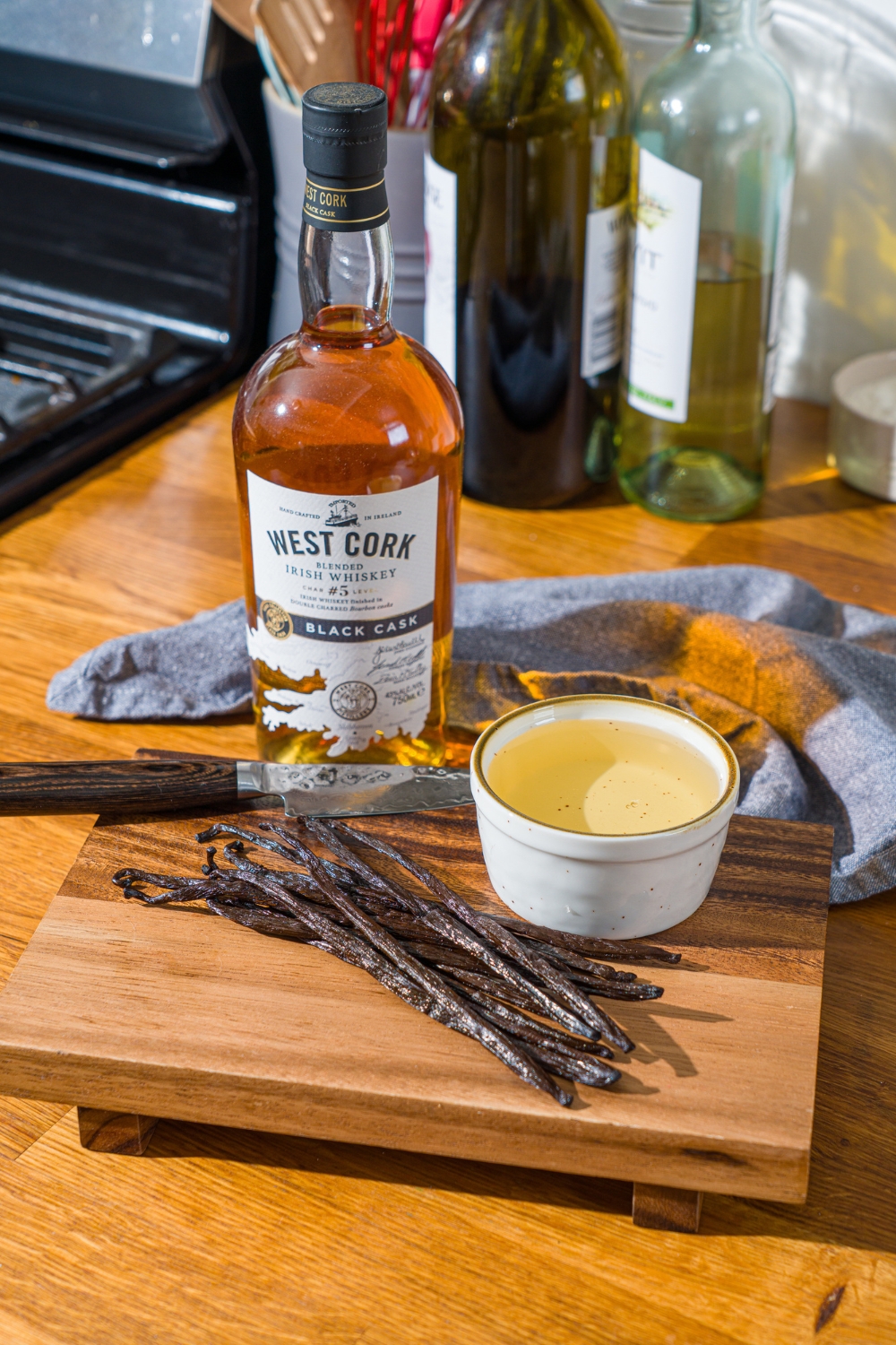A wooden board with ingredients to make homemade vanilla bean paste including whole vanilla beans, corn syrup, and a bottle of whiskey. The board is on a wooden counter with a blue cloth napkin.