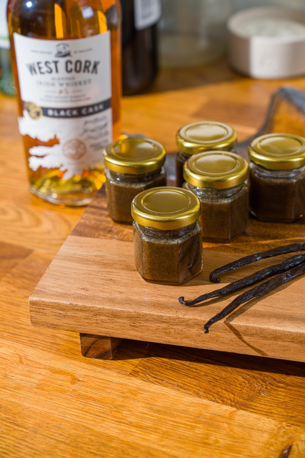A wooden board with several jars of homemade vanilla bean paste and whole vanilla beans. The board is on a wooden counter with a bottle of whiskey.
