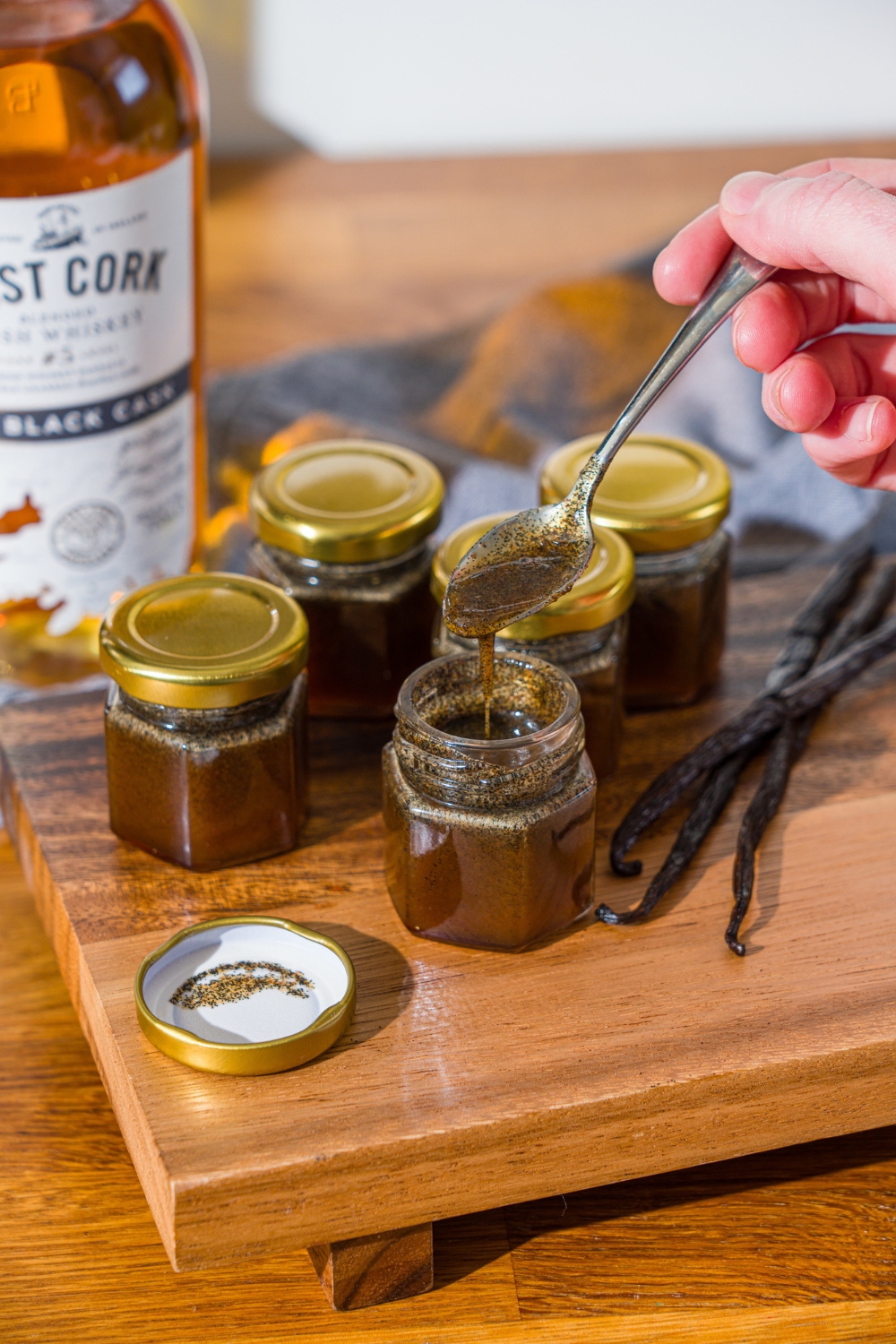 A wooden board with several jars of homemade vanilla bean paste and whole vanilla beans. A spoon is pouring the paste into one of the open jars. The board is on a wooden counter with a bottle of whiskey.