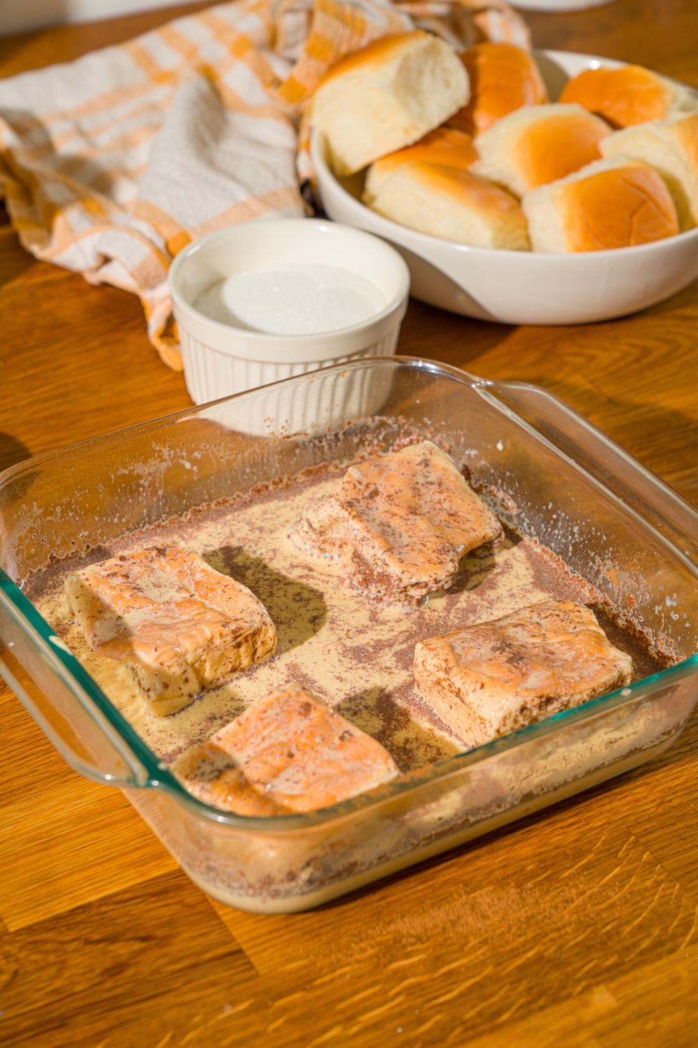 A baking dish with Hawaiian rolls soaking in french toast custard. The dish is on a wooden counter with a bowl of rolls and bowl of sugar.