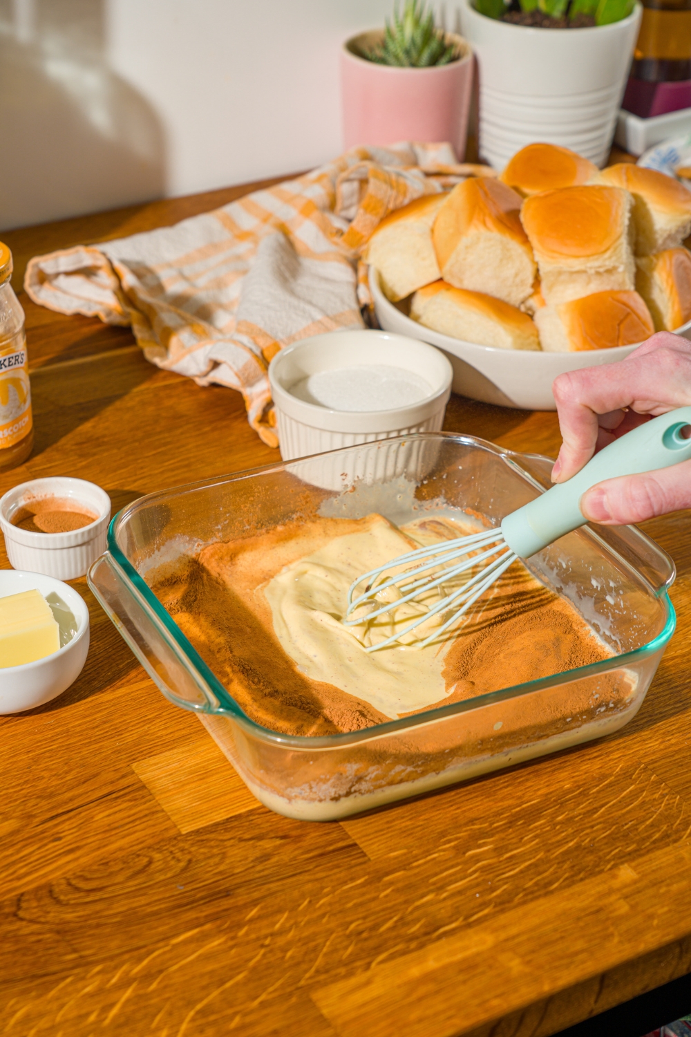 A whisk mixing a french toast custard mixture in a glass baking dish. The dish is on a wooden counter with a bowl of rolls and bowl of sugar.