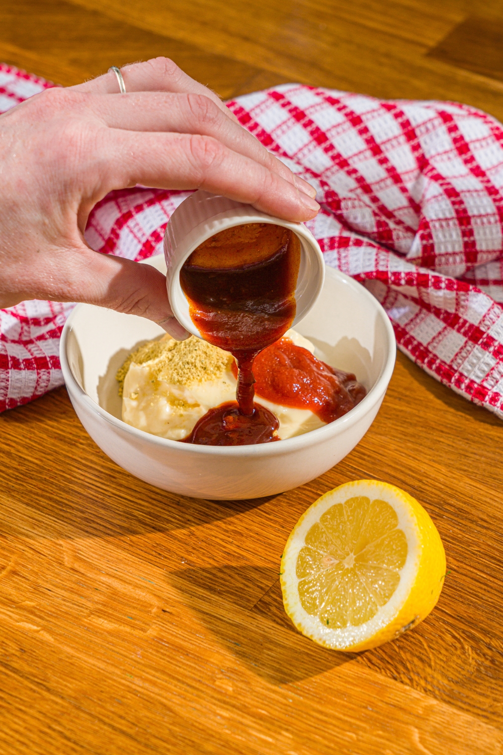 A hand pouring a small ramekin of chipotle sauce into a white bowl with ingredients to make comeback sauce including mayo, sriracha, and seasonings. The bowl is on a wooden counter with a lemon and red checkered napkin.