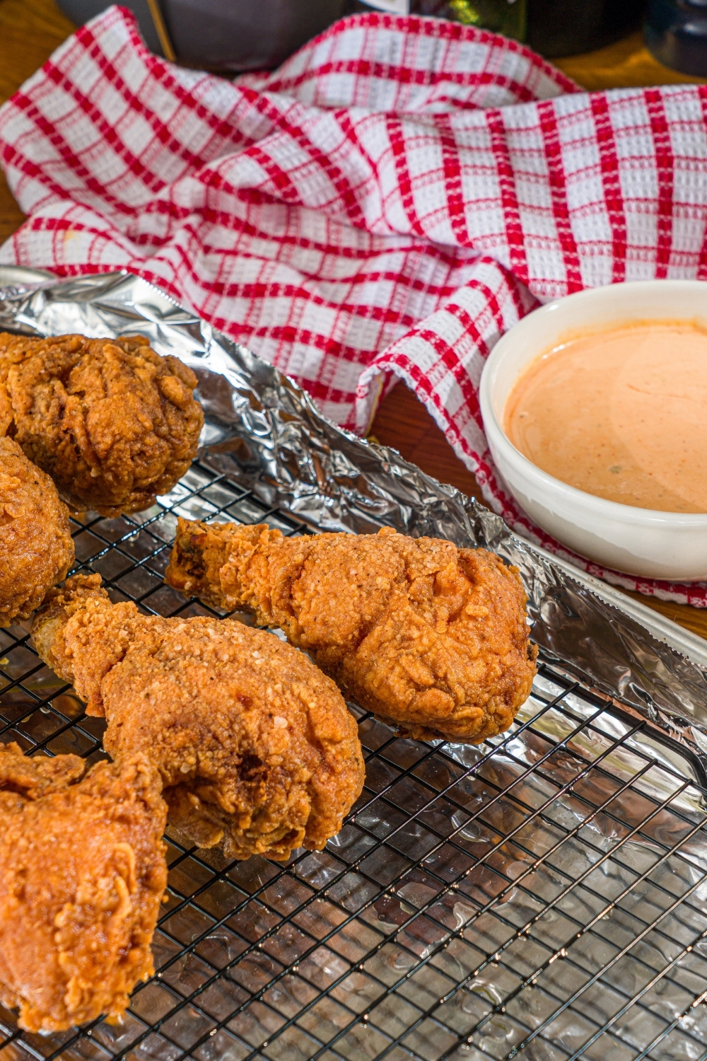 A baking sheet lined with foil topped with a drying rack with several pieces of copycat KFC fried chicken. The sheet is on a wooden counter with a bowl of mayo dipping sauce and red checkered napkin.