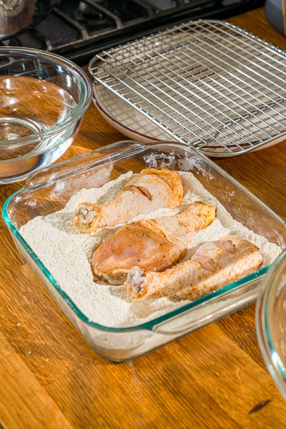 A glass baking dish with seasoned flour with a marinated chicken leg sitting in the flour. The dish is on a wooden counter.