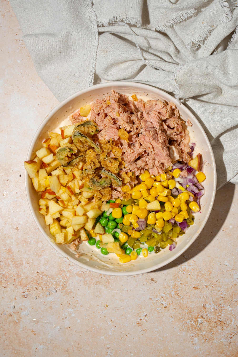 A large white bowl with ingredients to make Matthew McConaughey tuna salad including wasabi mayo dressing, tuna, corn, peas, apple, pickles, and jalapeño chips. The bowl is on a tan counter with a white cloth napkin.