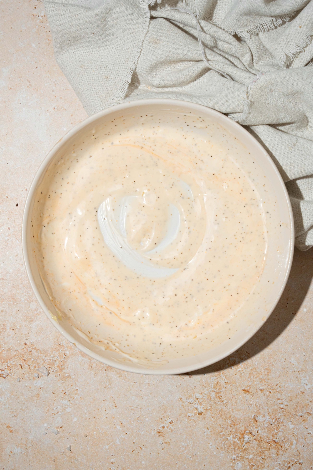 A large white bowl with whisked Matthew McConaughey tuna salad dressing including mayonnaise, wasabi paste, vinegar, and lemon juice. The bowl is on a tan counter with a white cloth napkin.