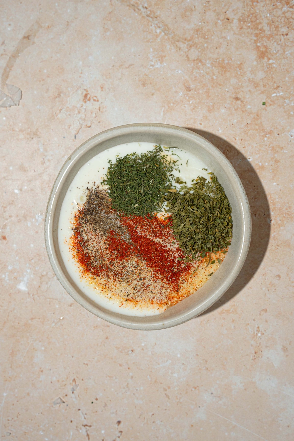 A white bowl with ingredients to make Popeyes blackened ranch including mayo, sour cream, buttermilk, and seasonings. The bowl is on a tan counter.