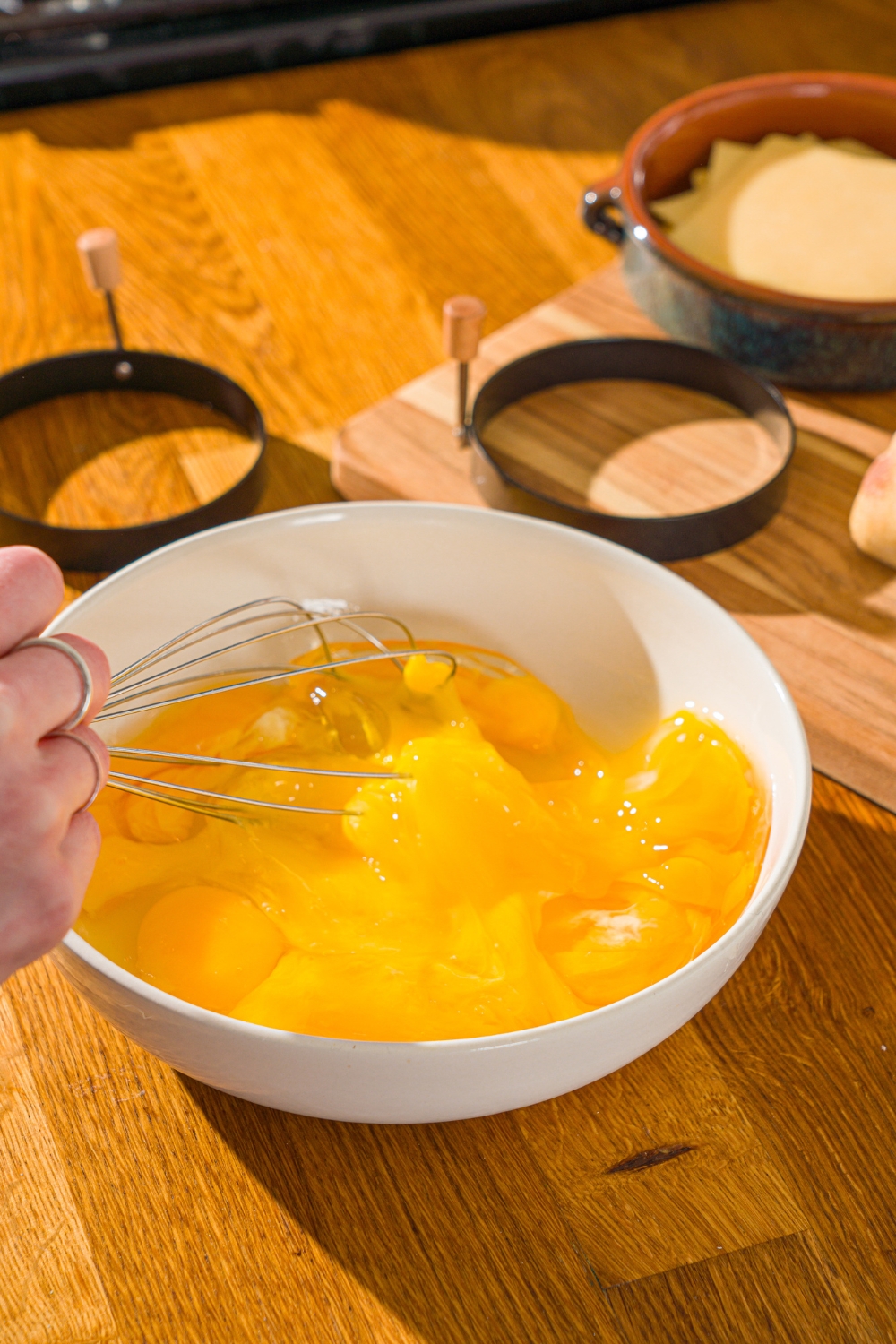 A large bowl with a whisk whisking eggs. The bowl is on a wooden counter with two round molds.