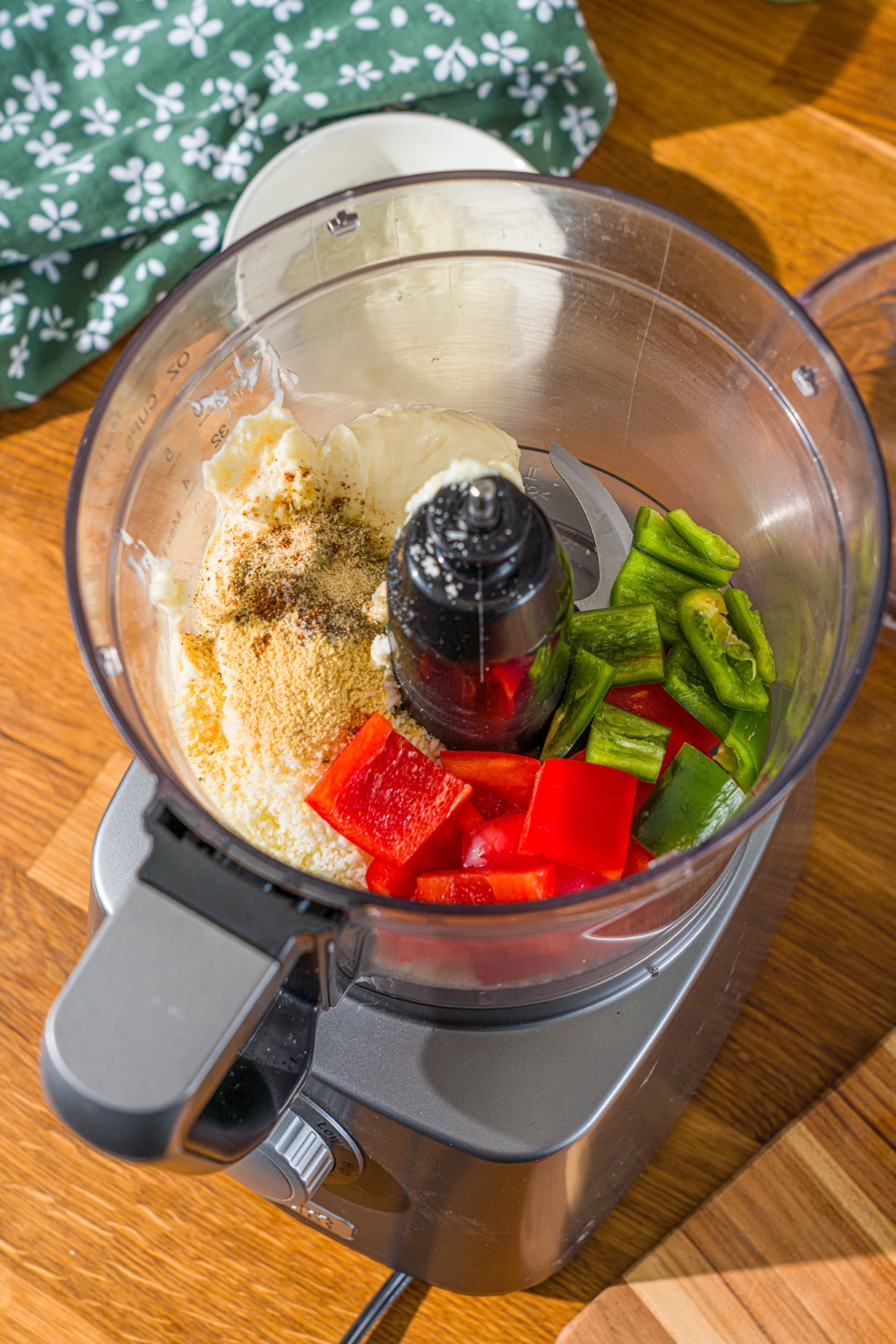 A food processor with ingredients to make Taco Bell baja sauce including red pepper, jalapeño, mayo, cotija cheese, and seasonings. The food processor is on a wooden counter with a green cloth napkin.