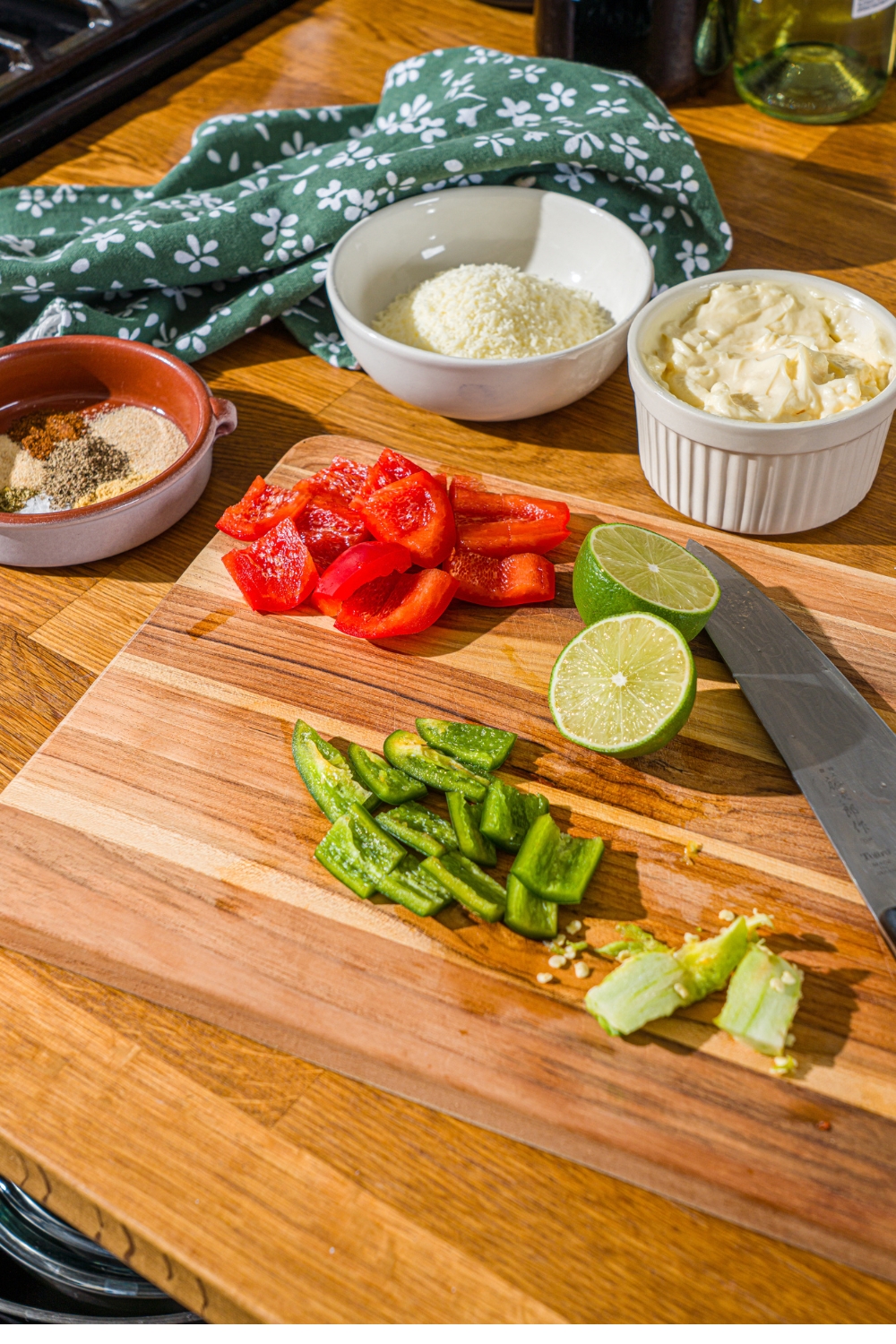 A wooden cutting board with a knife and chopped jalapeño and red pepper The board is on a wooden counter with bowls of ingredients.