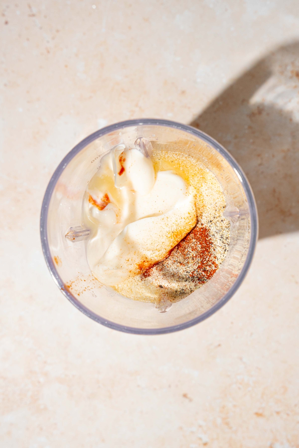 A blender jar with ingredients to make Taco Bell chipotle sauce including mayo, sour cream, chipotle peppers, and seasonings. The jar is on a tan counter.