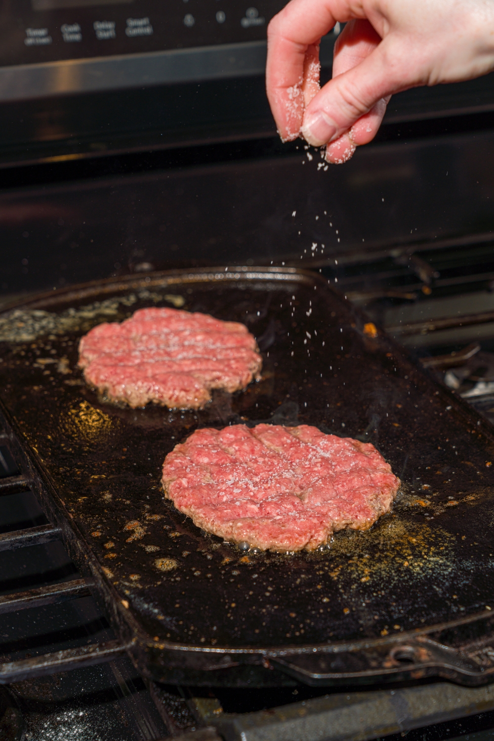 A stove with a hot griddle cooking two smashed beef patties while being seasoned with salt and pepper.