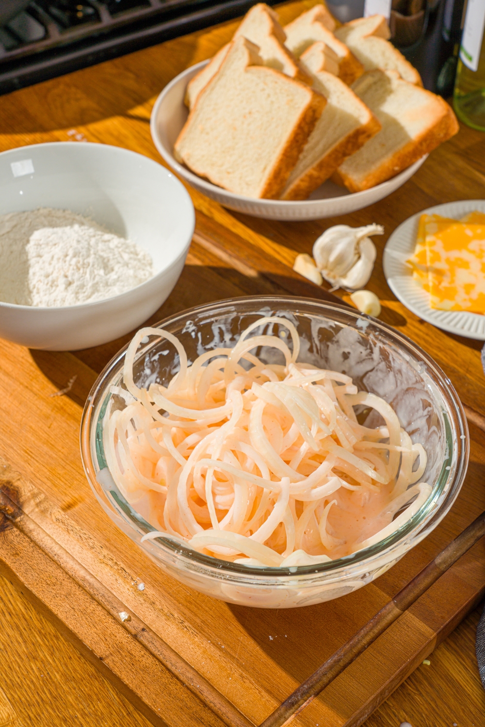 A glass bowl with sliced onions soaking in a buttermilk mixture. The bowl is on a wooden counter with a bowl of flour and sliced bread.