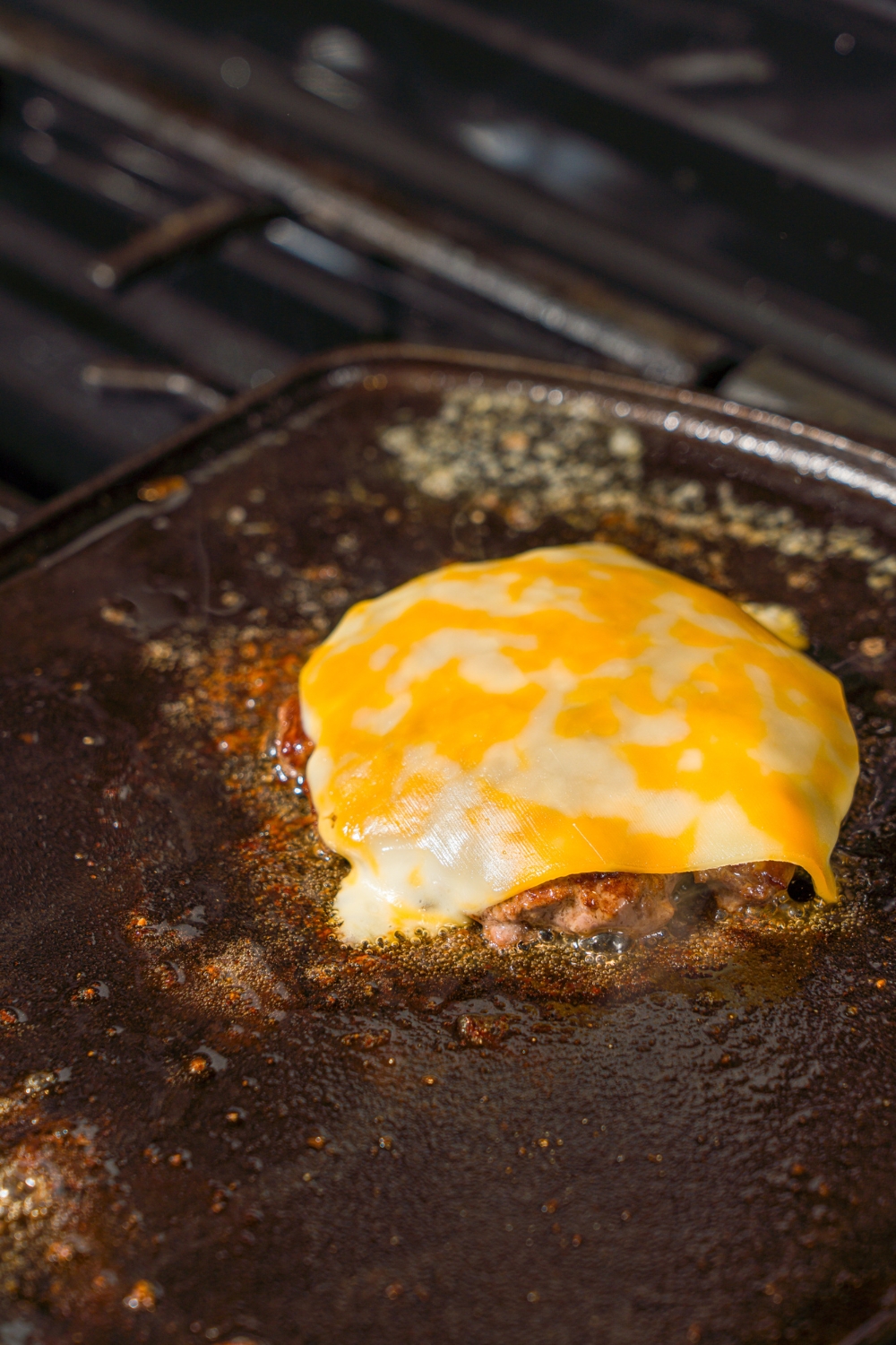 A stove with a hot griddle cooking a burger topped with cheese.