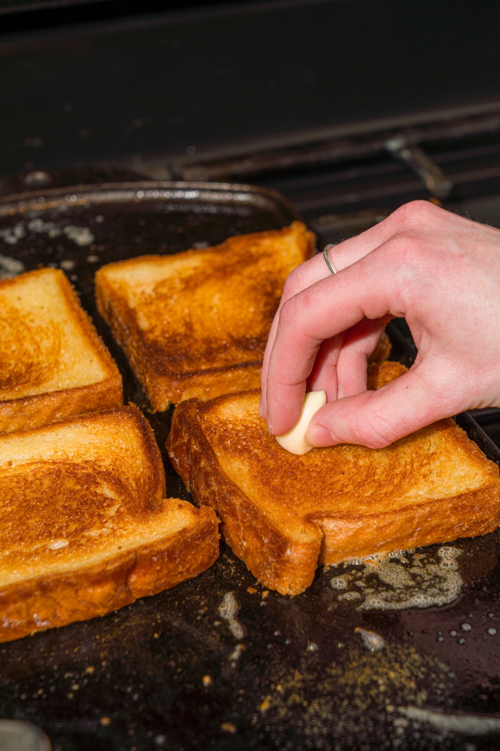 A stove with a hot griddle toasting four slices of bread. A hand is rubbing garlic on the bread.