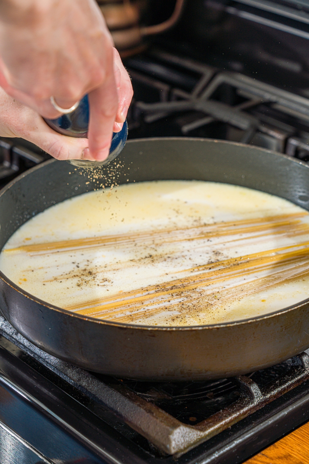 A skillet with a creamy garlic sauce base with uncooked spaghetti cooking on the stove. A hand is sprinkling pepper over the sauce.