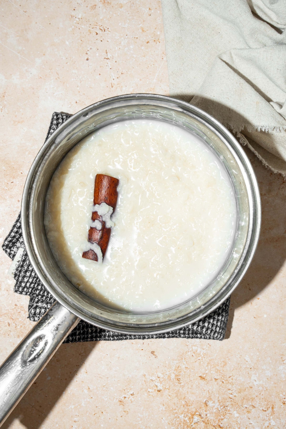 A pot with rice cooking in milk with a cinnamon stick. The pot is on a dish towel on a tan counter with a white cloth napkin.