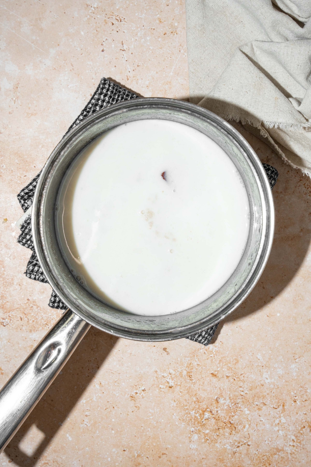 A pot with milk, water, rice, and cinnamon sticks. The pot is on a dish towel on a tan counter with a white cloth napkin.