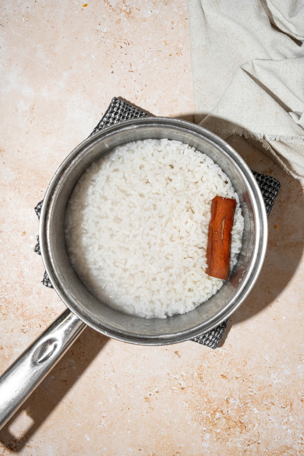 A pot with cooked rice with a cinnamon stick. The pot is on a dish towel on a tan counter.
