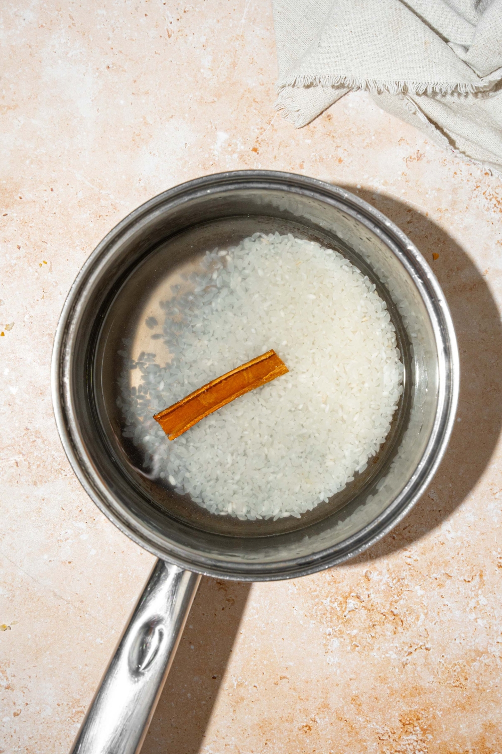 A pot with water, rice, and a cinnamon stick. The pot is on a dish towel on a tan counter.
