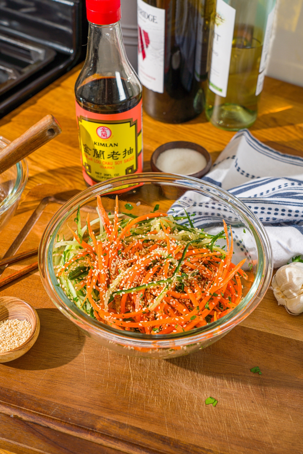 A glass bowl with shredded carrots and cucumber tossed in dressing and topped with sesame seeds. The bowl is on a wooden cutting board.