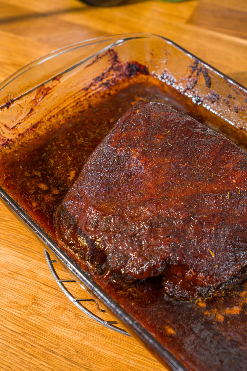 A glass baking dish with oven baked beef brisket rubbed with seasonings and bbq sauce. The dish is on a wooden counter.