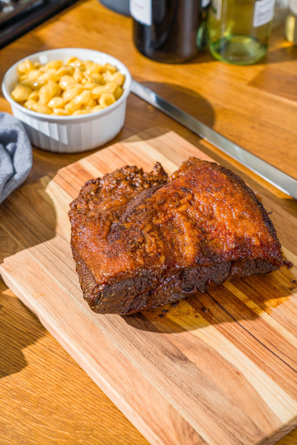 A wooden cutting board with oven baked beef brisket rubbed with seasonings and bbq sauce, and a bowl of mac and cheese. The board is on a wooden counter