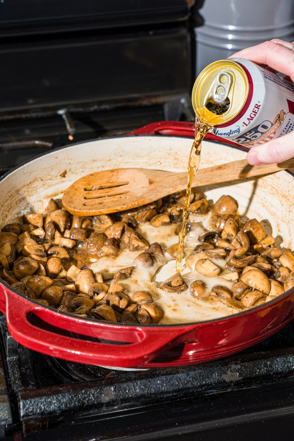 A cast iron skillet with roasted mushrooms. A can of beer is being poured into the skillet.